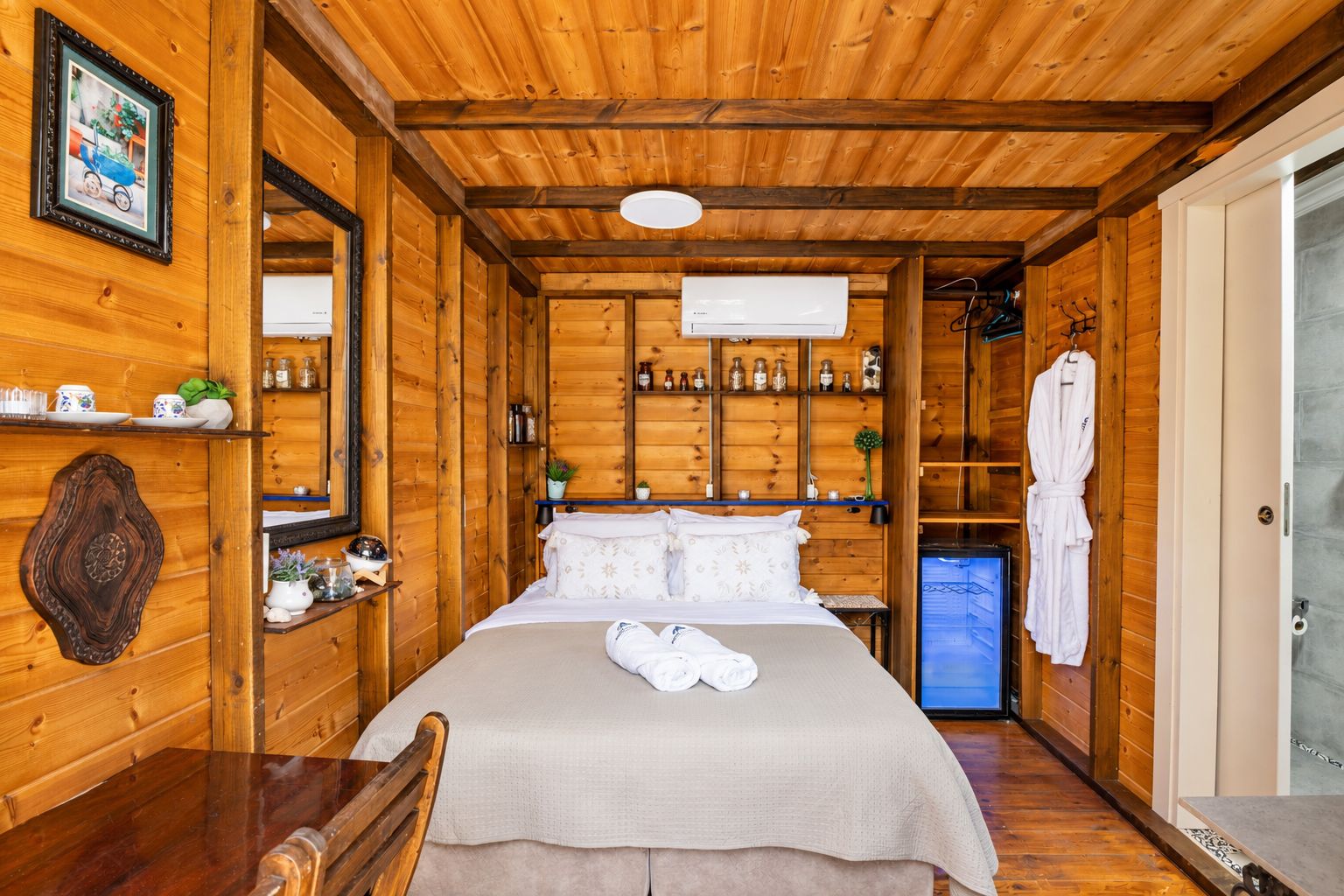 A wooden cabin room with a bed, shelves with toiletries, a robe hanging, and a small refrigerator.