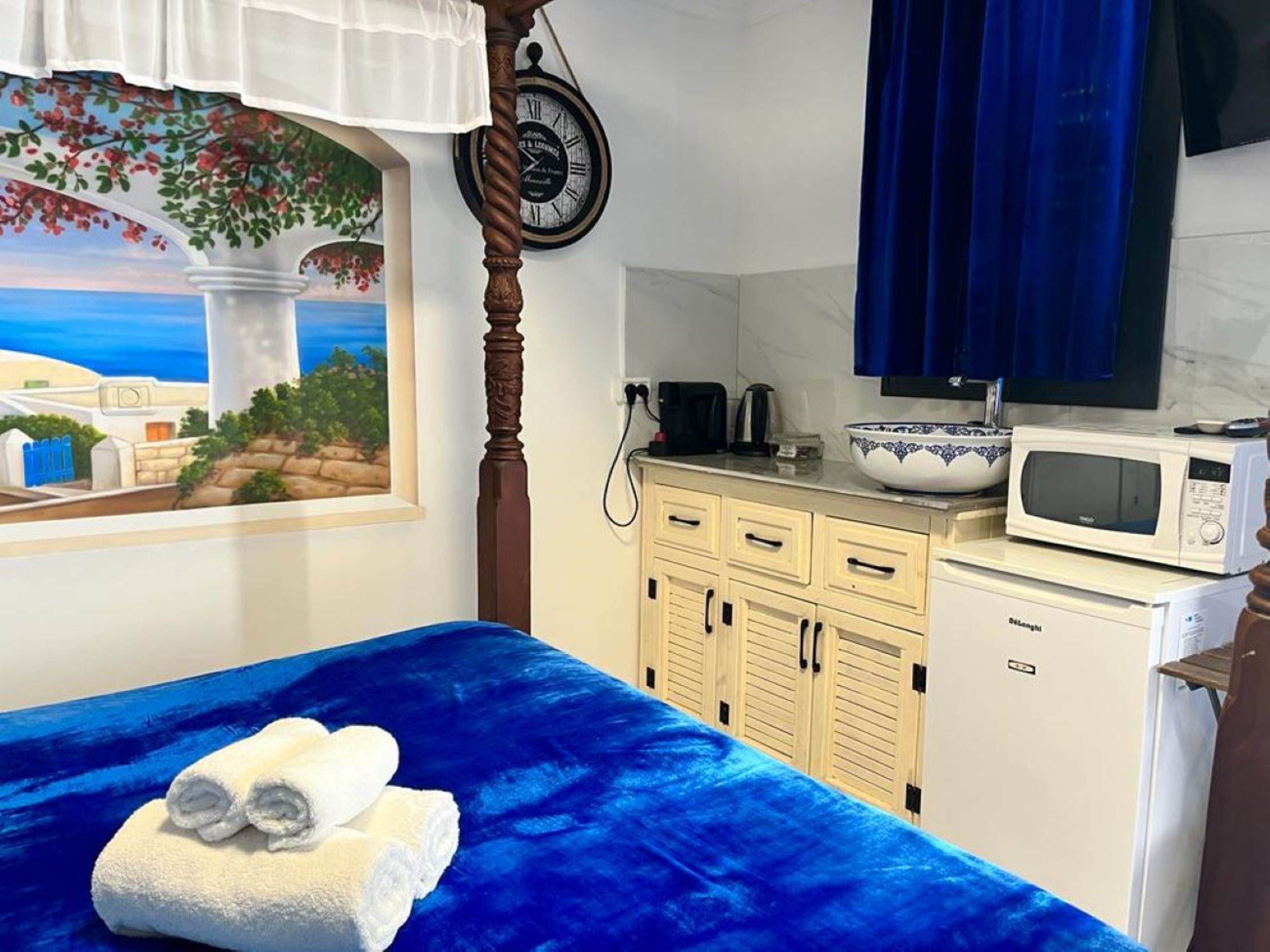 A bedroom with a blue bedspread, a painting of a coastal scene, a wooden bed frame, a white bedside table with a microwave, a sink, and a cabinet.
