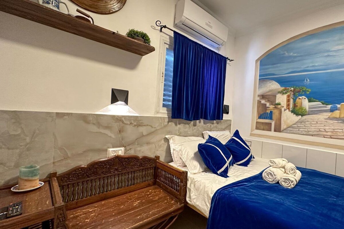 Un dormitorio con una cama, un banco de madera, un estante en la pared con plantas, un cuadro de una escena costera y una ventana con cortinas azules.
