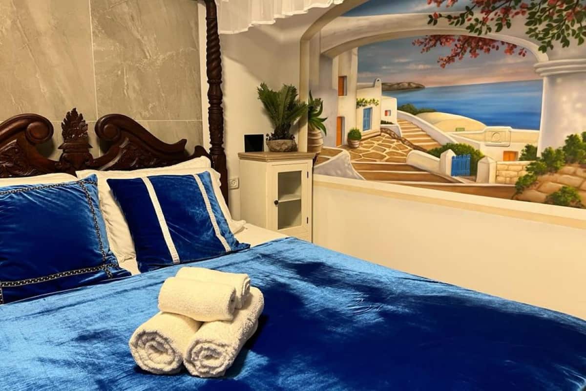 A bedroom with a bed, pillows, and a view of a coastal scene through an arched window.