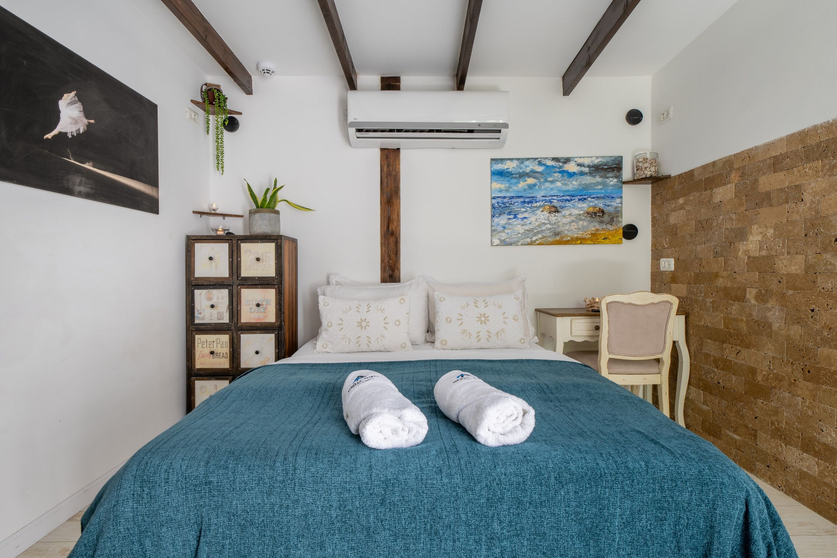 A bedroom with a bed, a chest of drawers, a desk, a chair, a painting, and an air conditioning unit.