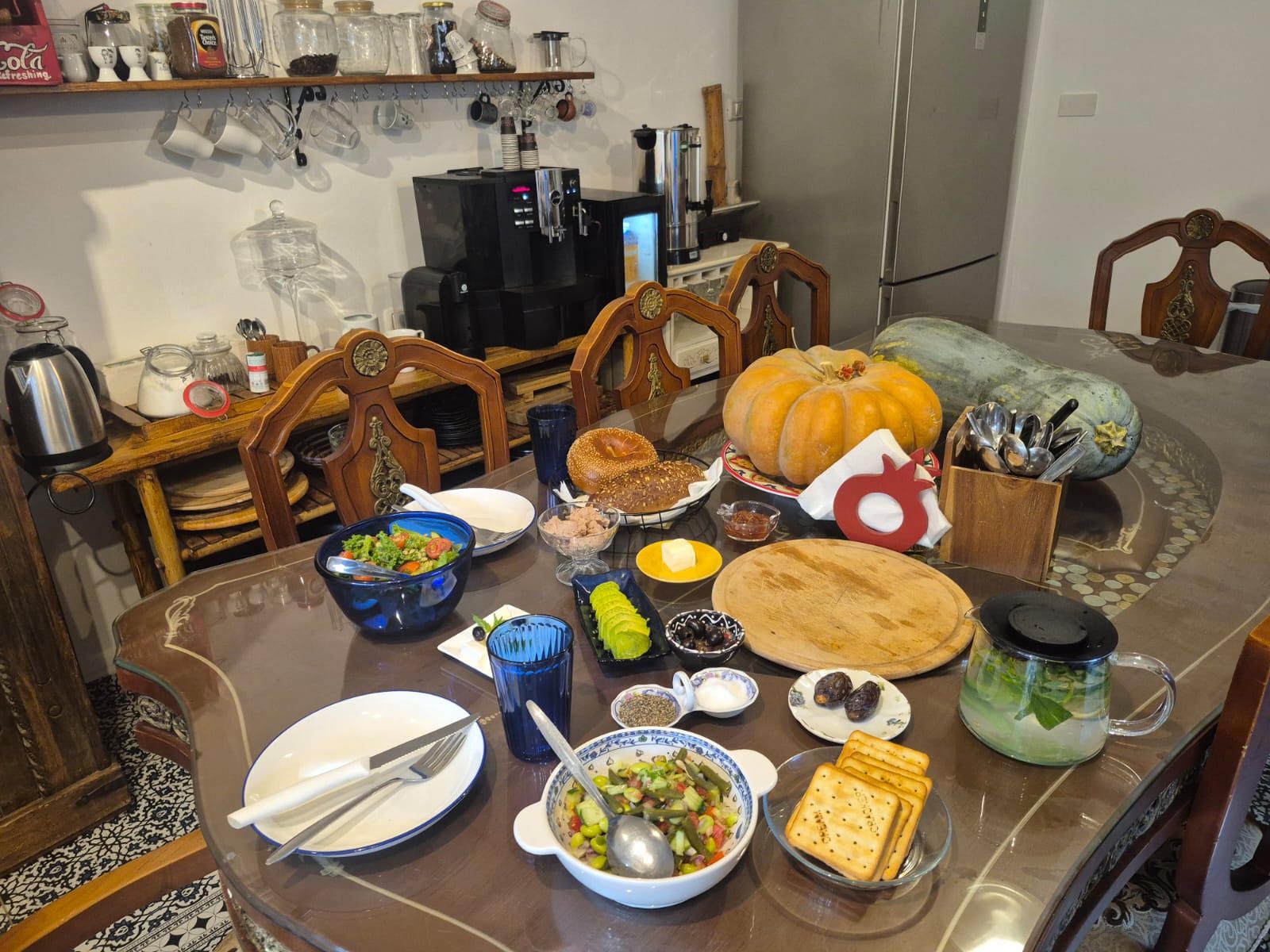 Una mesa de comedor dispuesta con diferentes platos, una máquina de café y un centro de mesa con calabazas.