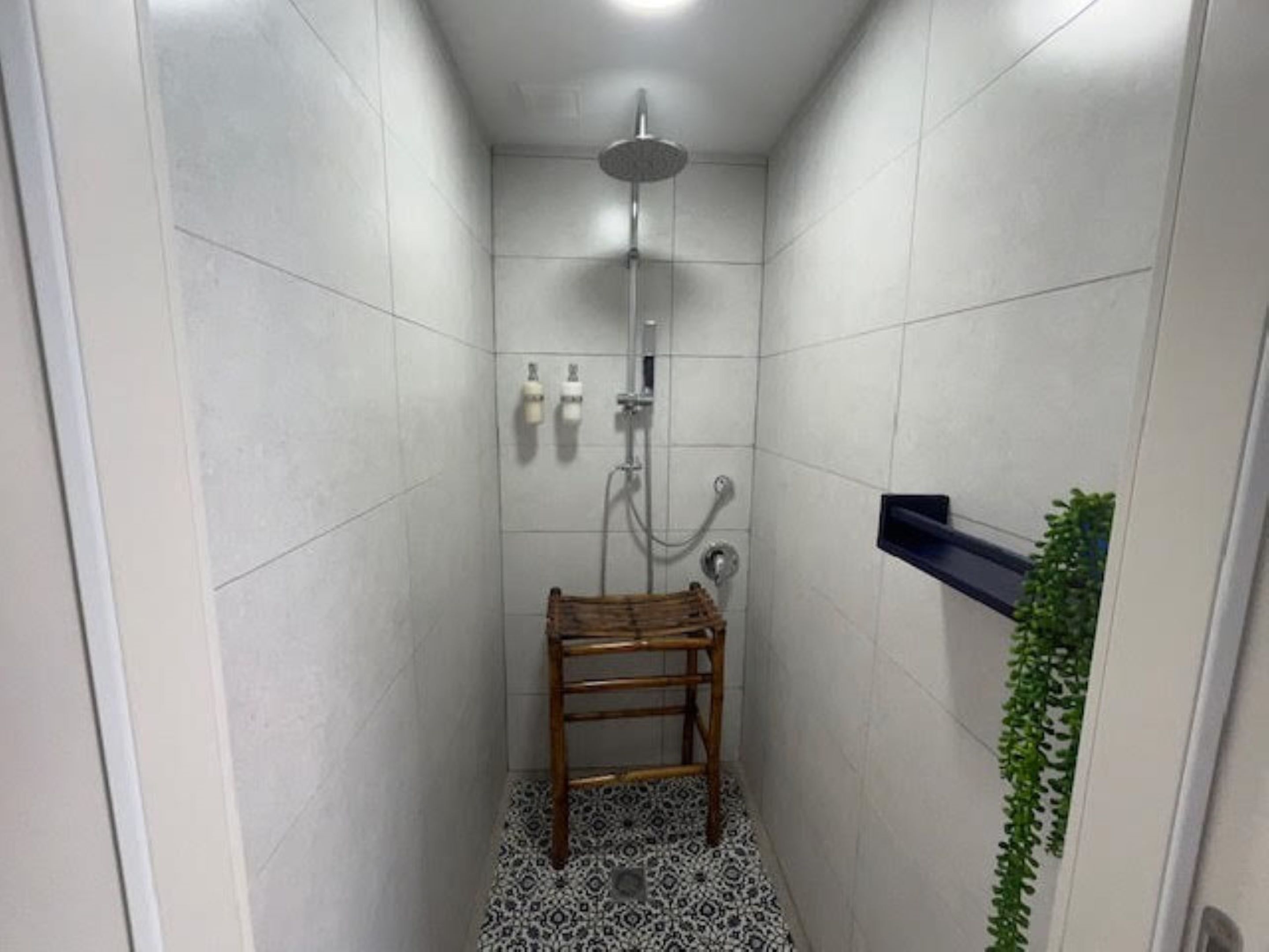 A small, tiled shower room with a wooden stool, a wall-mounted shelf, and a potted plant.