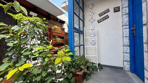Una entrada de edificio con una puerta azul, un estante de madera con plantas y una pared con letreros numerados.