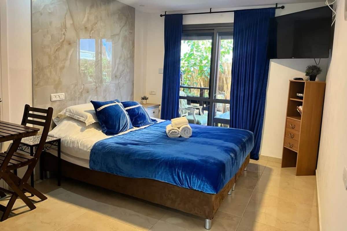 A bedroom with a bed, a wooden chair, a small table, a TV, and a window with curtains.