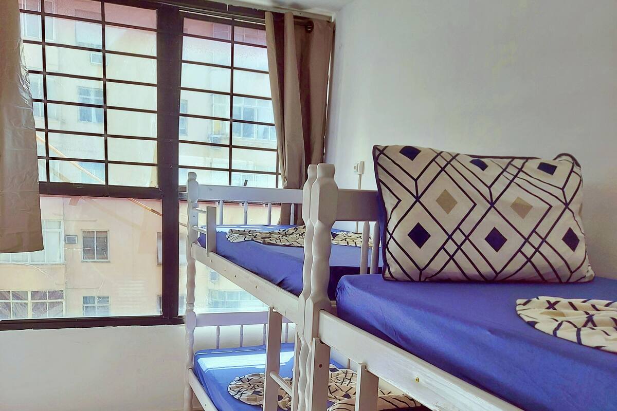 A dormitory room with bunk beds, a window with curtains, and a view of buildings outside.