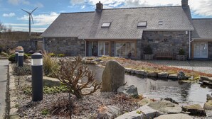 A stone house with a pond, a wind turbine, and a clear sky.