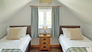 A bedroom with two beds, a window with curtains, a lamp, and a wooden nightstand. - 2 bedroom accommodation in Prion, near Denbigh (Prion, near Denbigh)