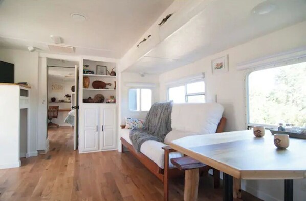 A cozy room with a wooden floor, a white sofa with a grey throw, a wooden table, and a window with a view of trees.