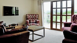 A living room with a sofa, armchair, and a table. There is a television mounted on the wall and a large window with a view of greenery. - 5 bedroom accommodation in Aviemore (Aviemore)
