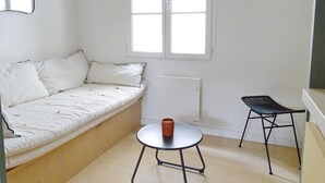 Een kleine, lichte kamer met een witte bank, een klein rond tafeltje met een kopje en een zwarte stoel.