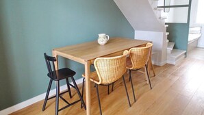 Een eetruimte met een houten tafel en twee rieten stoelen, een witte kruik op tafel en een trap op de achtergrond.