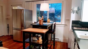 A compact kitchen with a refrigerator, a wooden table, and a window with a view of houses. - Sunlit Craftsman Home | Steps to Cafes and restaurants | 10 mn to downtown (Vancouver)