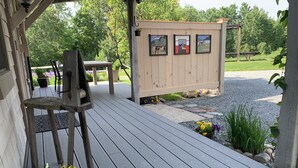 A wooden deck with a bench and potted flowers, a small wooden structure with framed pictures, and a gravel path leading to a grassy area. - Pristine Custom Cottage, Grand Piano, Massage Studio on Site (Leicester)