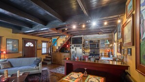 A cozy living room with a sofa, coffee table, and a dining area with a table set for two. A kitchen with modern appliances is visible in the background. - Pristine Custom Cottage, Grand Piano, Massage Studio on Site (Leicester)