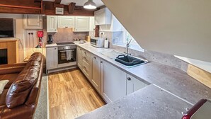 A compact kitchen with a built-in oven, microwave, and sink. There is a wooden floor, a brown leather sofa, and a window providing natural light.