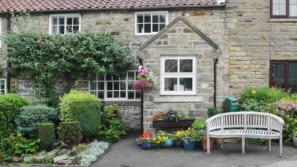 A stone house with a tiled roof, a bench, and potted plants in the garden.