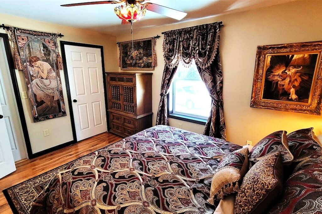 A bedroom with a patterned bedspread, a ceiling fan, a window with curtains, and a door with a mirror.