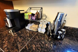 A coffee station with a coffee maker, a basket of coffee pods, and a set of stainless steel cups and utensils.