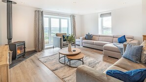 A living room with a fireplace, a sofa, a coffee table, and a view through large windows. - Hazelgrove Lodge Barn (Laneshawbridge)