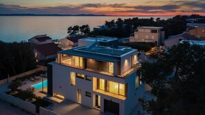 Ett modernt hus i två våningar med en takterrass, en swimmingpool och utsikt över havet vid solnedgången.