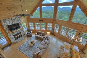 A spacious living room with a stone fireplace, large windows, and a wooden ceiling.