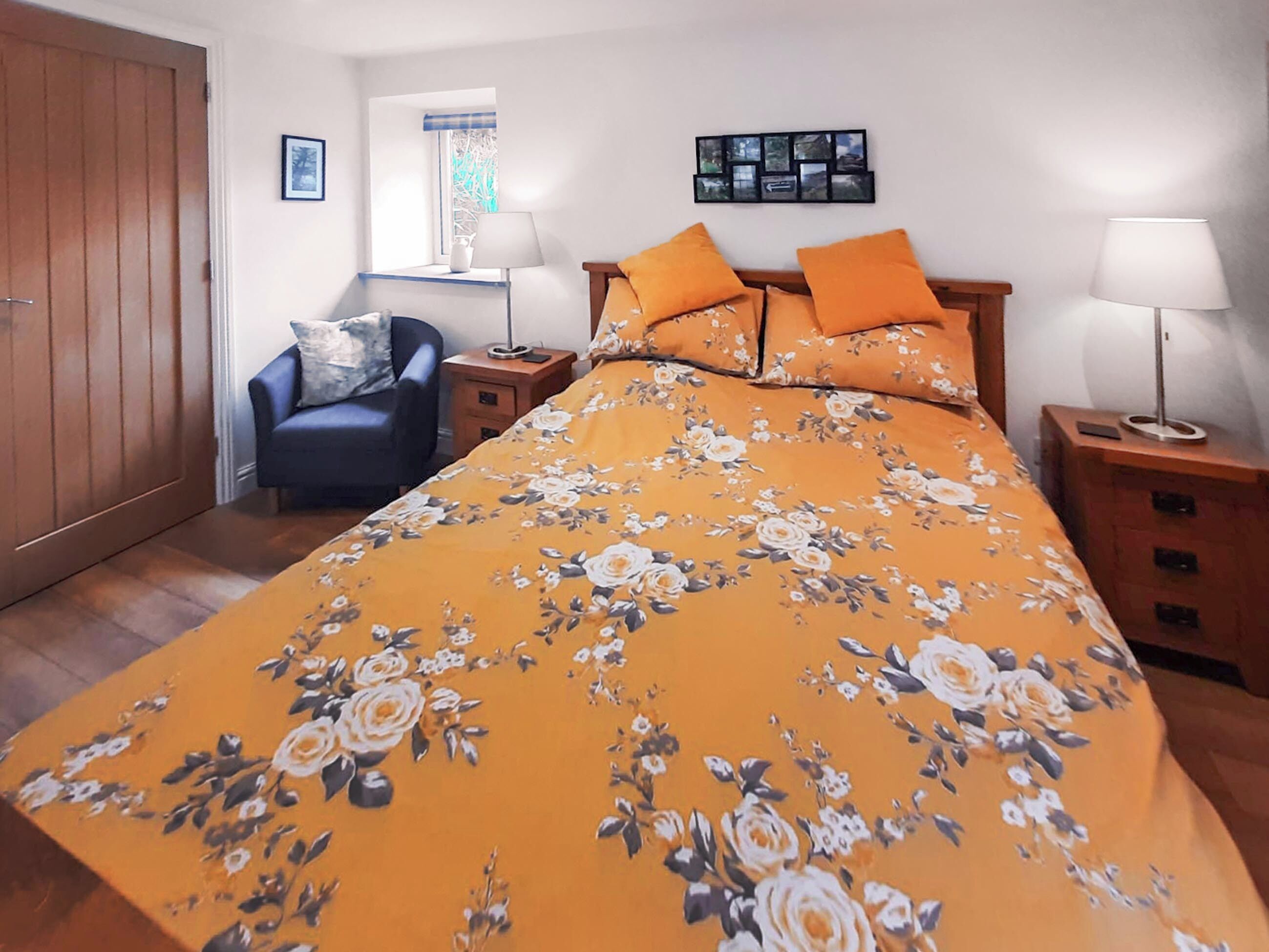 A bedroom with a floral bedspread, two bedside lamps, a nightstand, a chair, a window with a view, and a wooden wardrobe.