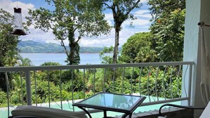 A balcony with a glass table and two chairs, overlooking a pool and lush greenery.