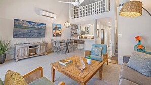 A spacious living area with a wooden coffee table, a sofa, and a dining table. There is a ceiling fan, a kitchen in the background, and a staircase leading to an upper level.