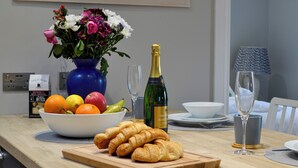 A dining table with a vase of flowers, a bottle of champagne, a bowl of fruit, and croissants. - Grange Loft - UK31594 (Dundee)