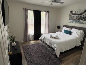 Un dormitorio con una cama, una mesita de noche, una planta, una ventana con cortinas y un ventilador de techo.