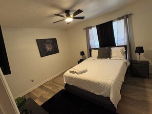 Quarto com cama, ventilador de teto, mesa de cabeceira com abajur, janela com cortinas e um quadro na parede.