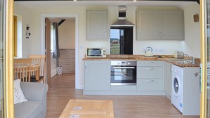 A compact kitchen with a microwave, oven, and washing machine. There is a wooden table and a sofa in the living area. - 1 bedroom accommodation in Alkham, near Dover (Alkham, near Dover)