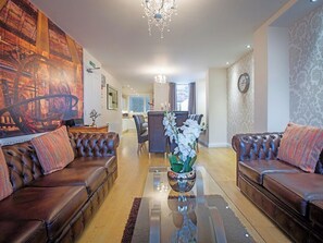 A spacious living room with leather sofas, a glass coffee table, and a decorative vase with white flowers. A large mural and a clock adorn the walls.