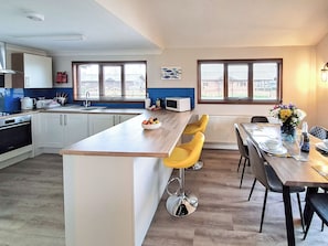 A modern kitchen with a central island, bar stools, a dining table with chairs, and a view through the windows. - Perfect water’s edge holiday accommodation, set in a quiet marina. (Brundall)