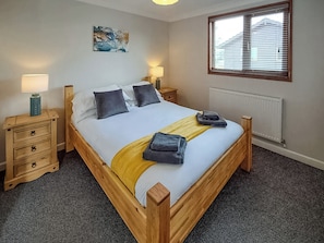 A bedroom with a wooden bed, two bedside tables, a radiator, and a window with blinds. - Perfect water’s edge holiday accommodation, set in a quiet marina. (Brundall)