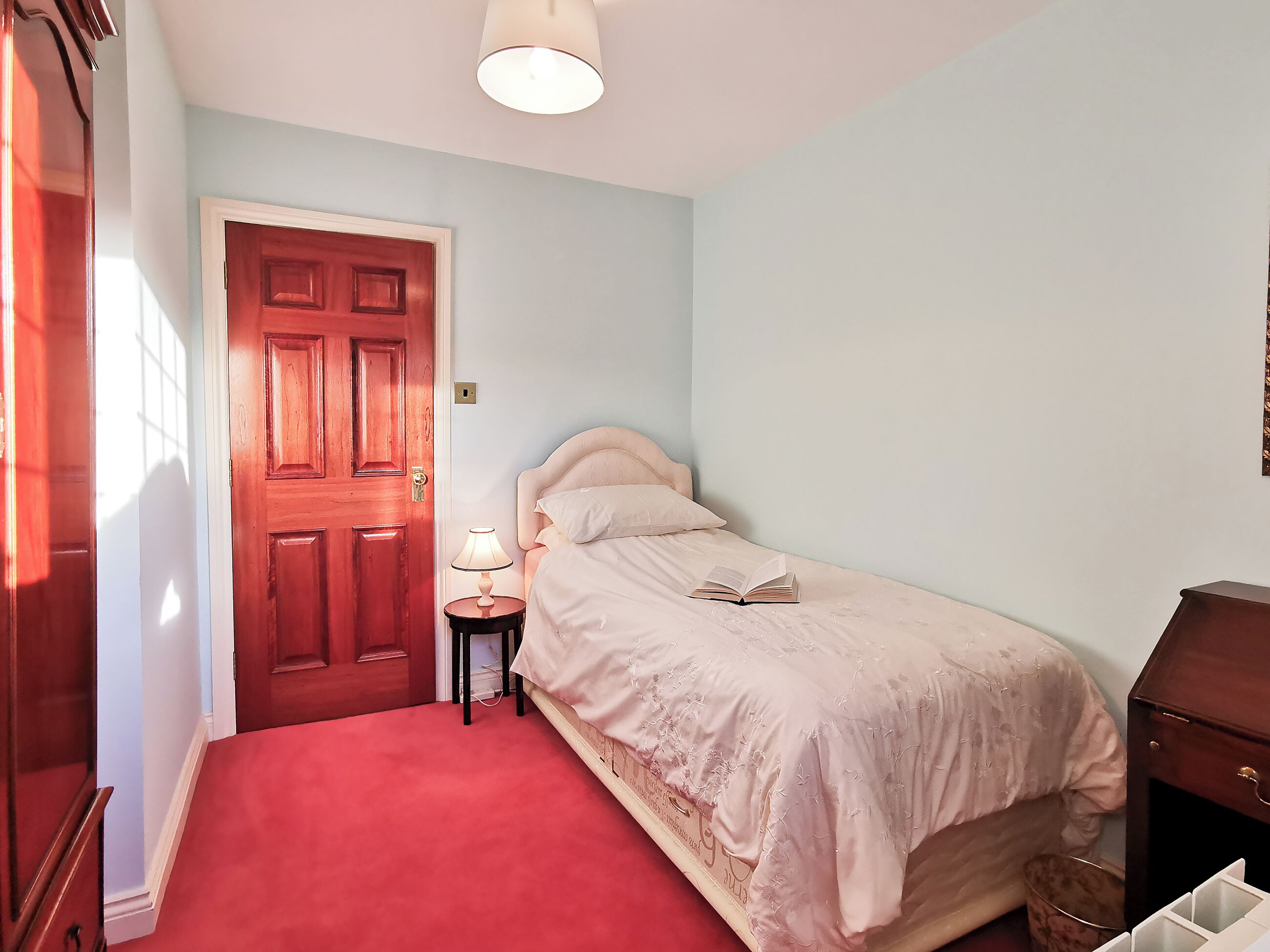 Un dormitorio con puerta de madera, lámpara de noche, cama con un libro y una mesita pequeña.