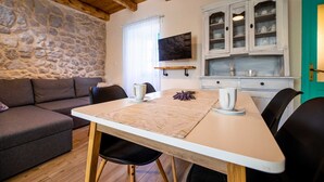 A cozy living room with a stone wall, a wooden ceiling, a TV mounted on the wall, a dining table with chairs, and a kitchen area with cabinets. - Charming holiday house Antica with private swimming pool (Rasopasno (Krk))