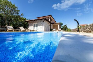 A swimming pool with lounge chairs, a stone house with a tiled roof, and a clear blue sky.
