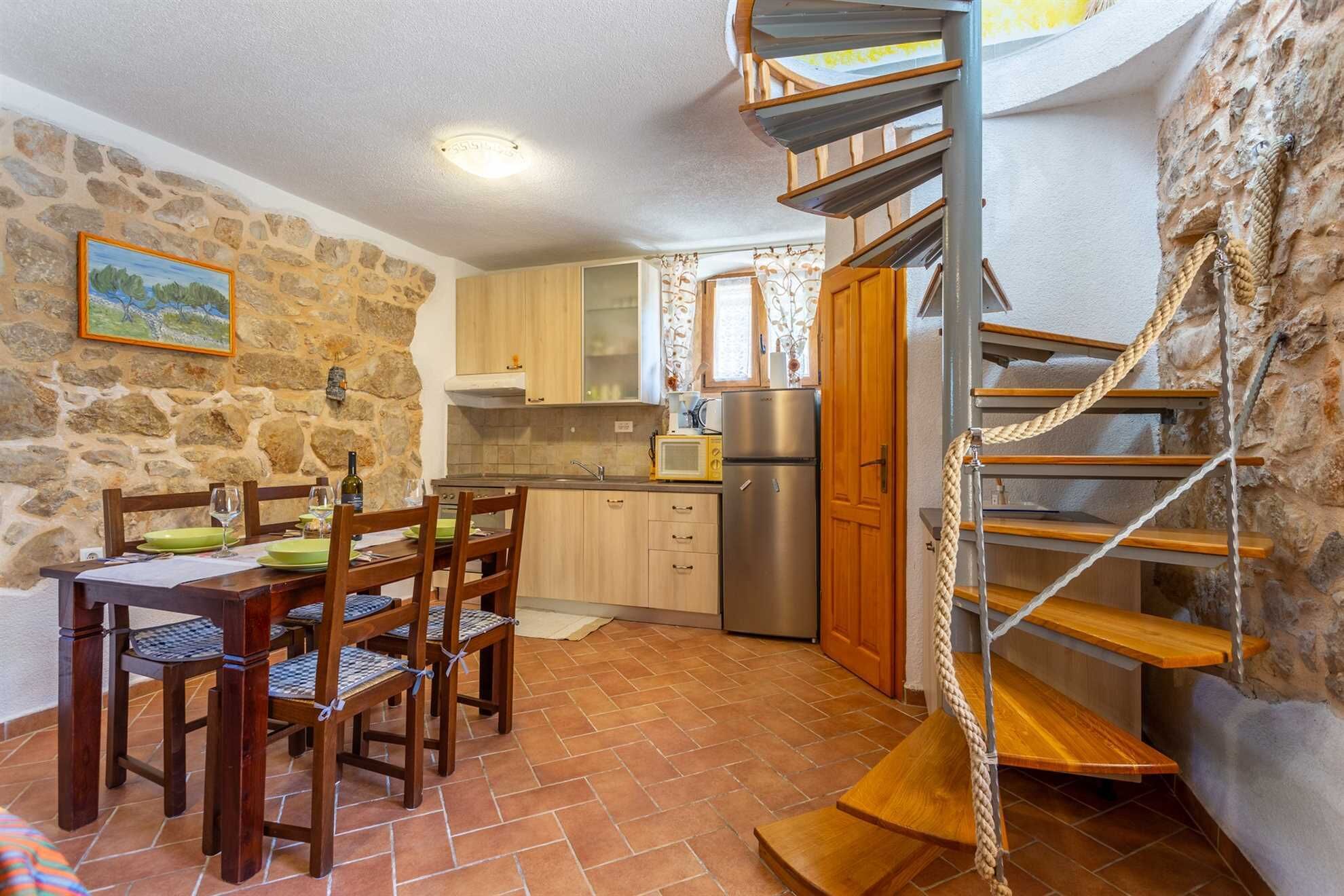 A compact kitchen with a stone wall, wooden cabinets, a dining area with a table and chairs, and a spiral staircase.