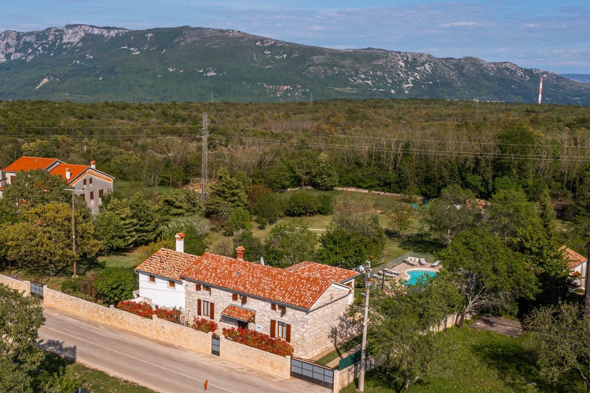 Une maison en pierre avec un toit recouvert de tuiles rouges, entourée de verdure et adossée à une montagne.