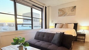 A modern hotel room with a large window, a bed with a white duvet, a dark sofa, a small table with a plant, and a nightstand with a lamp.