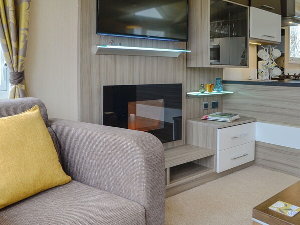 A modern living room with a flat-screen TV, a sofa with a yellow pillow, a small side table, and a kitchen area with a microwave and glassware.