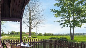 Un espace extérieur abrité, doté d’une terrasse en bois, d’une table avec des chaises et offrant une vue sur un champ verdoyant bordé d’arbres.