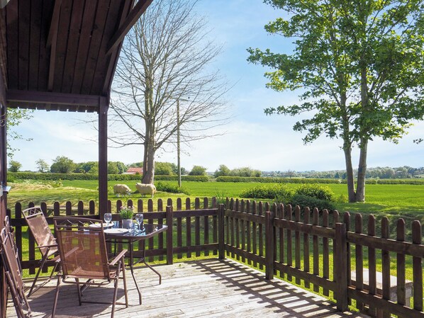 A covered outdoor area with a wooden deck, a table with chairs, and a view of a green field with trees.