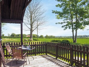 Un espacio al aire libre techado con una terraza de madera, una mesa con sillas y vistas a un campo verde con árboles.