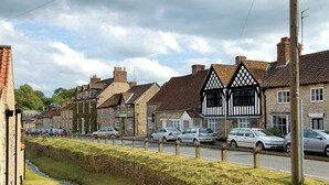Une rangée de maisons traditionnelles en pierre, avec une structure en bois apparent, une rue pavée et des voitures garées.