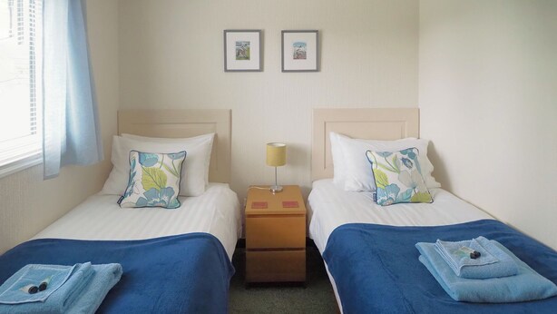 Une chambre d’hôtel avec deux lits, chacun recouvert d’une couverture et doté d’un oreiller bleu, une table de chevet avec une lampe, et deux tableaux encadrés accrochés au mur.