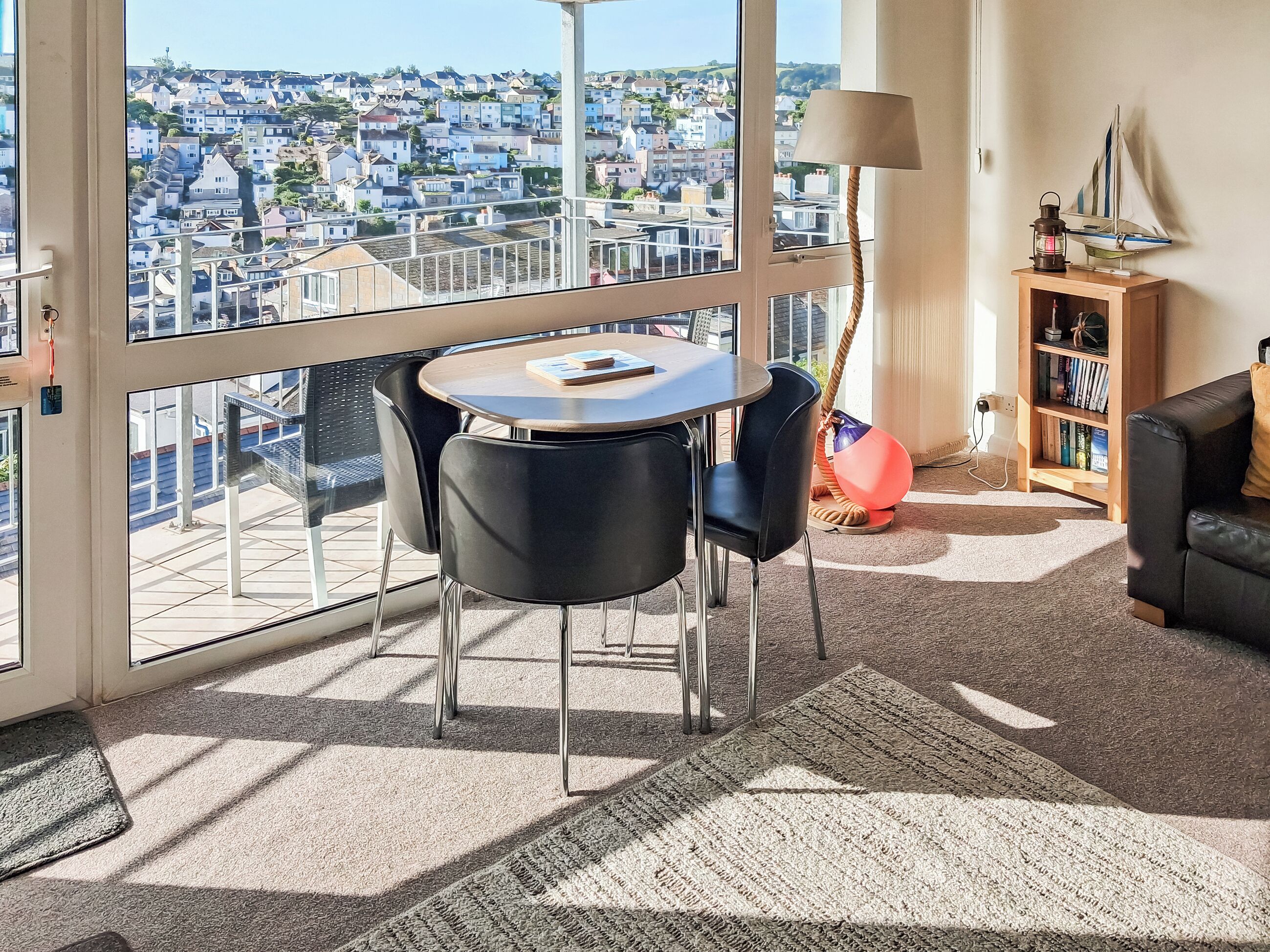 A modern living room with a large window, a round table, black chairs, a lamp, a bookshelf, and a view of a residential area.