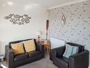A living room with two black leather sofas, a wooden side table with a lamp, a clock, and a radiator. - Upper Deck - 5 Linden Court (Brixham)